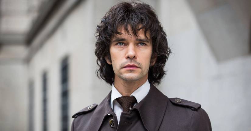 Norman standing in a black wrap around trench coat while staring off at something in A Very English Scandal.