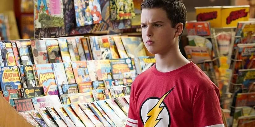 Iain Armitage as Sheldon wears a Flash T-shirt and looks stern as he stands beside a shelf of comic books