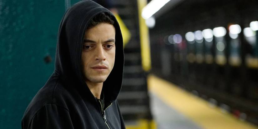 Rami Malek as Elliot, standing on an NYC subway platform, wearing a black hoodie, in Mr. Robot