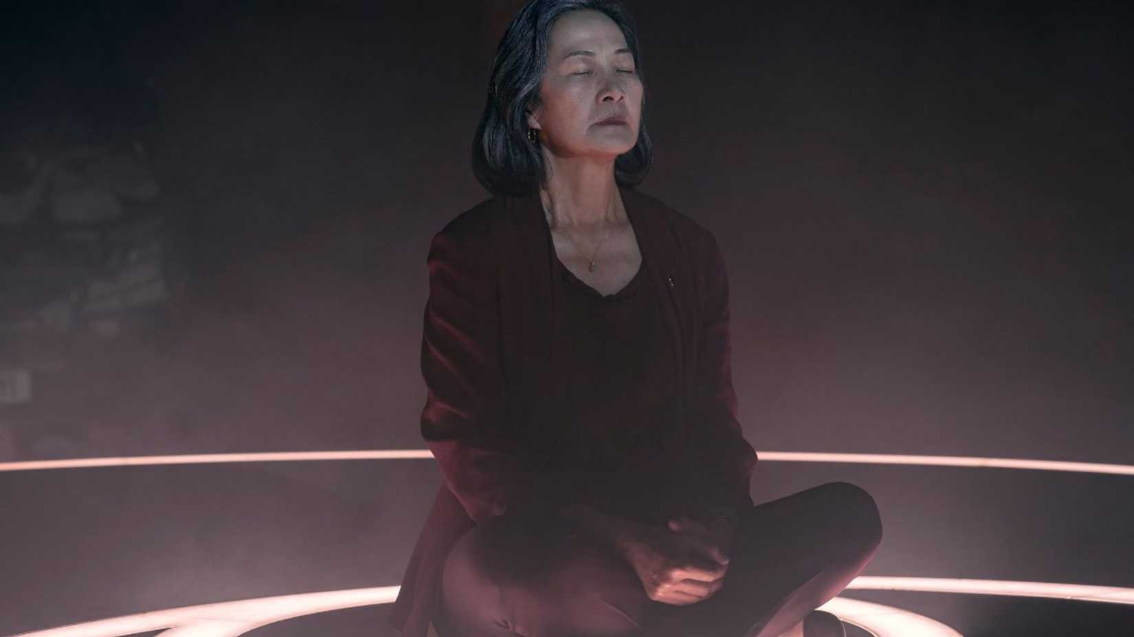 Розалинда Чао в роли доктора Е Вэньцзе сидит, скрестив ноги, в темной комнате с закрытыми глазами в «3 проблемах с телом».
