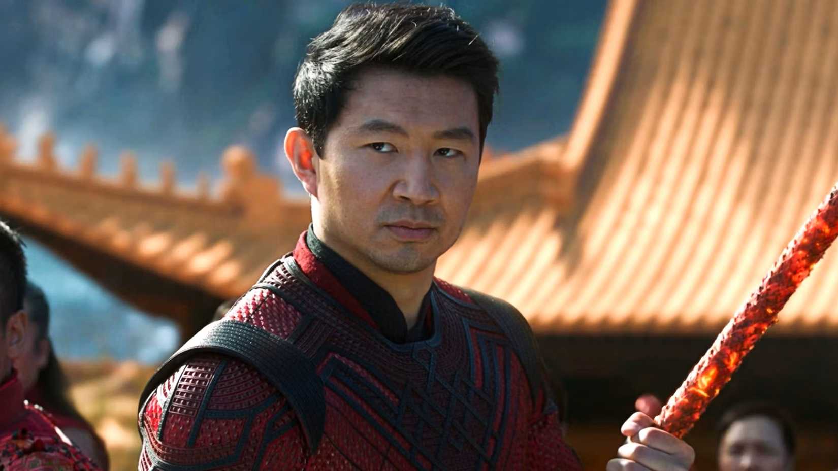 Simu Liu as Shang-Chi standing near a temple holding a weapon