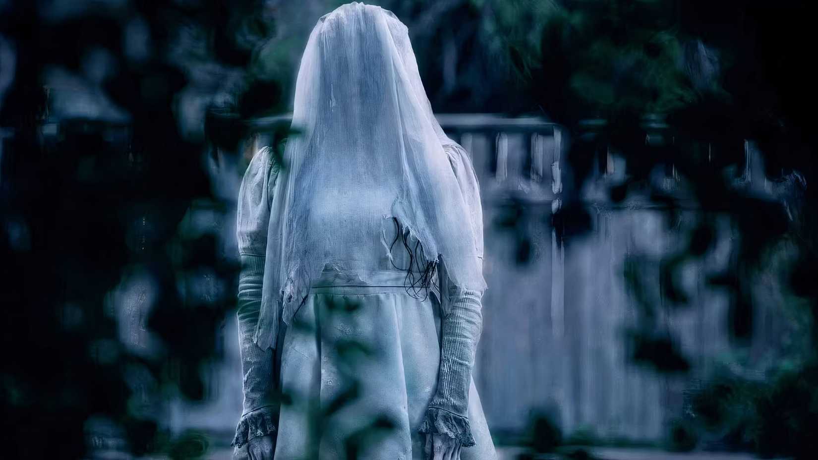 Marisol Ramirez as The Weeping Woman, wearing a white wedding dress and face covered by a veil, standing in a wooded area in The Curse of La Llorona