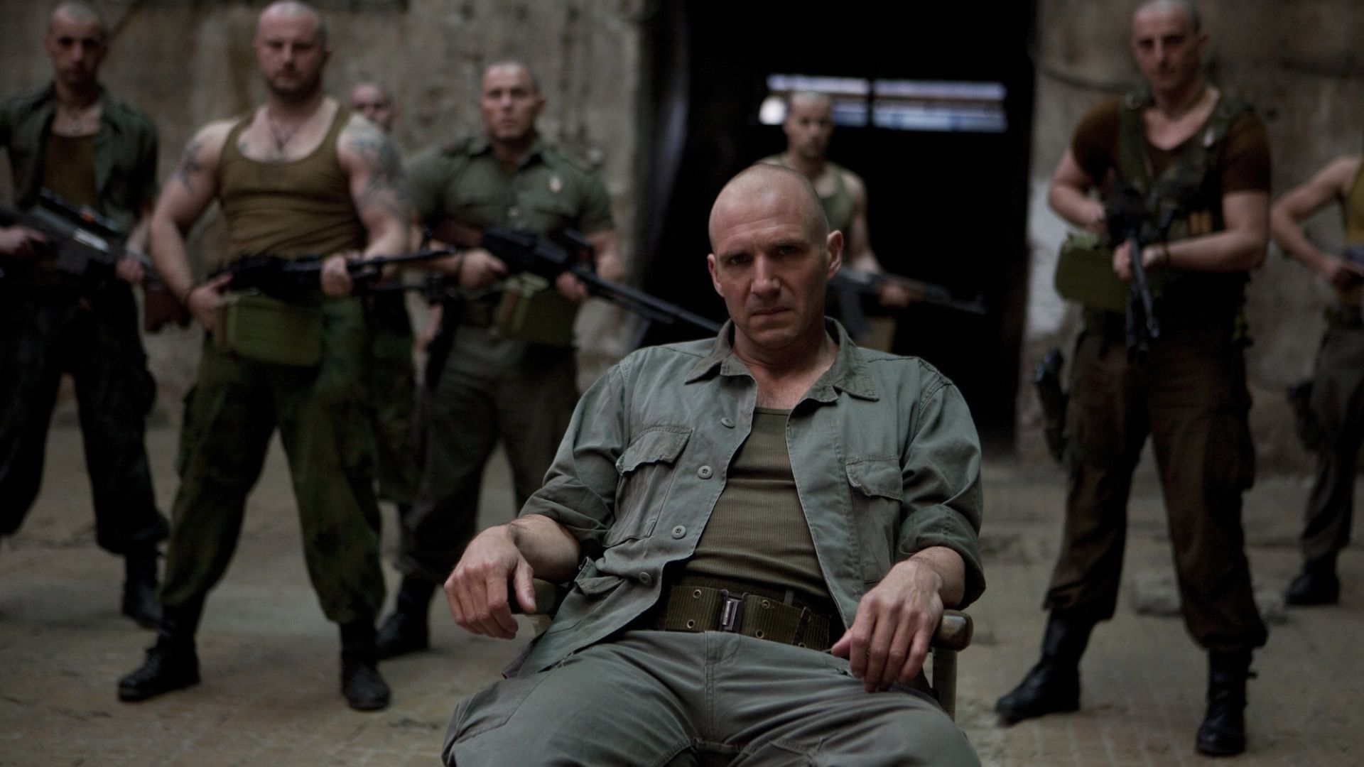 Ralph Fiennes site on a chair with thugs behind him in Coriolanus (2011)