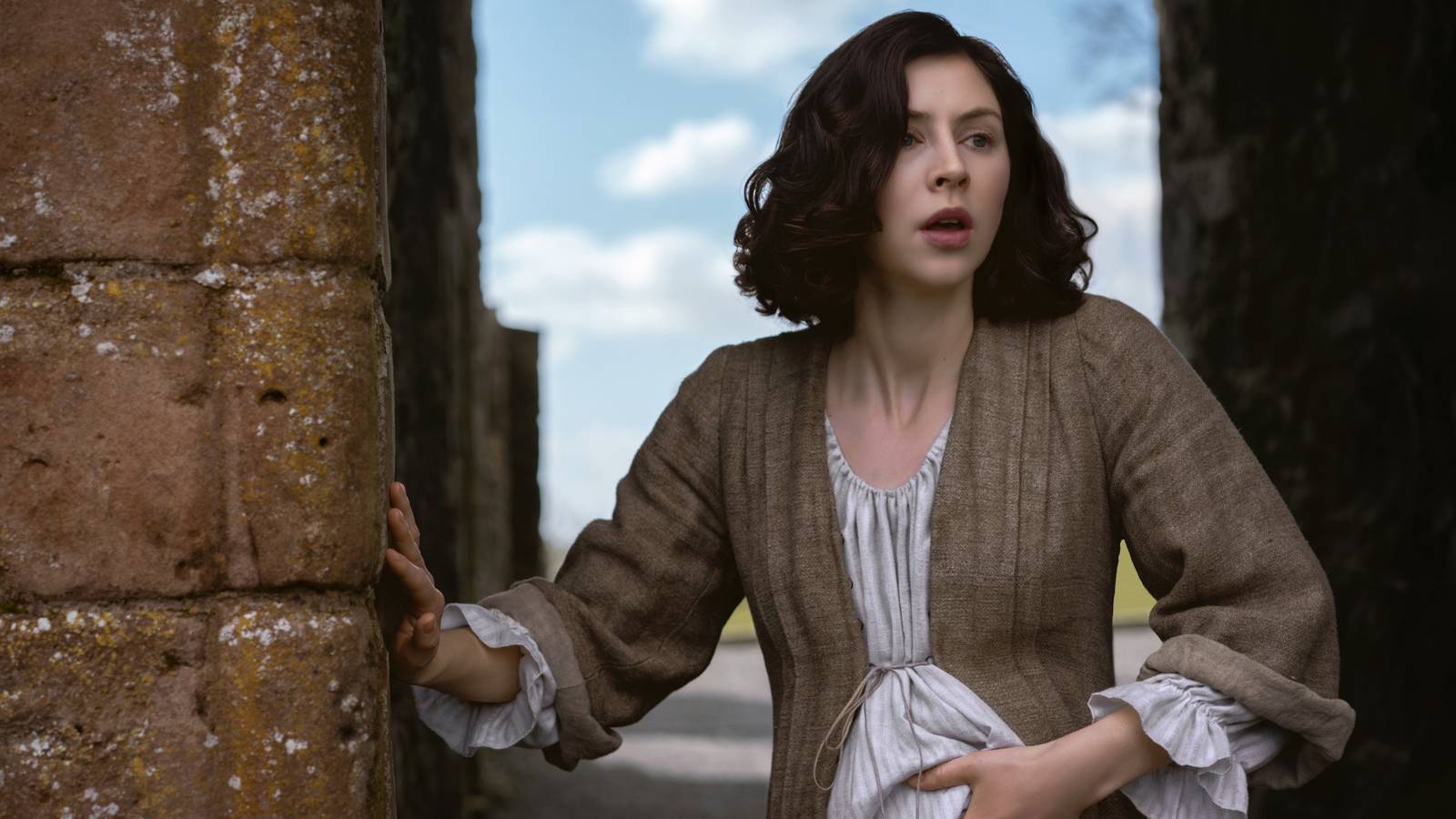 Hermione Corfield in Outlander: Blood of My Blood holding her stomach standing near a wall