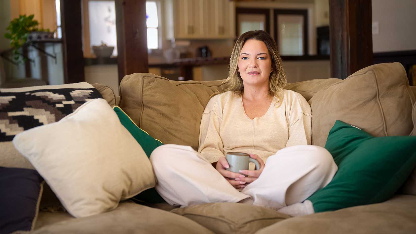 Bridget Phillips sitting cross-legged on a couch holding a coffee mug in Love Con Revenge.