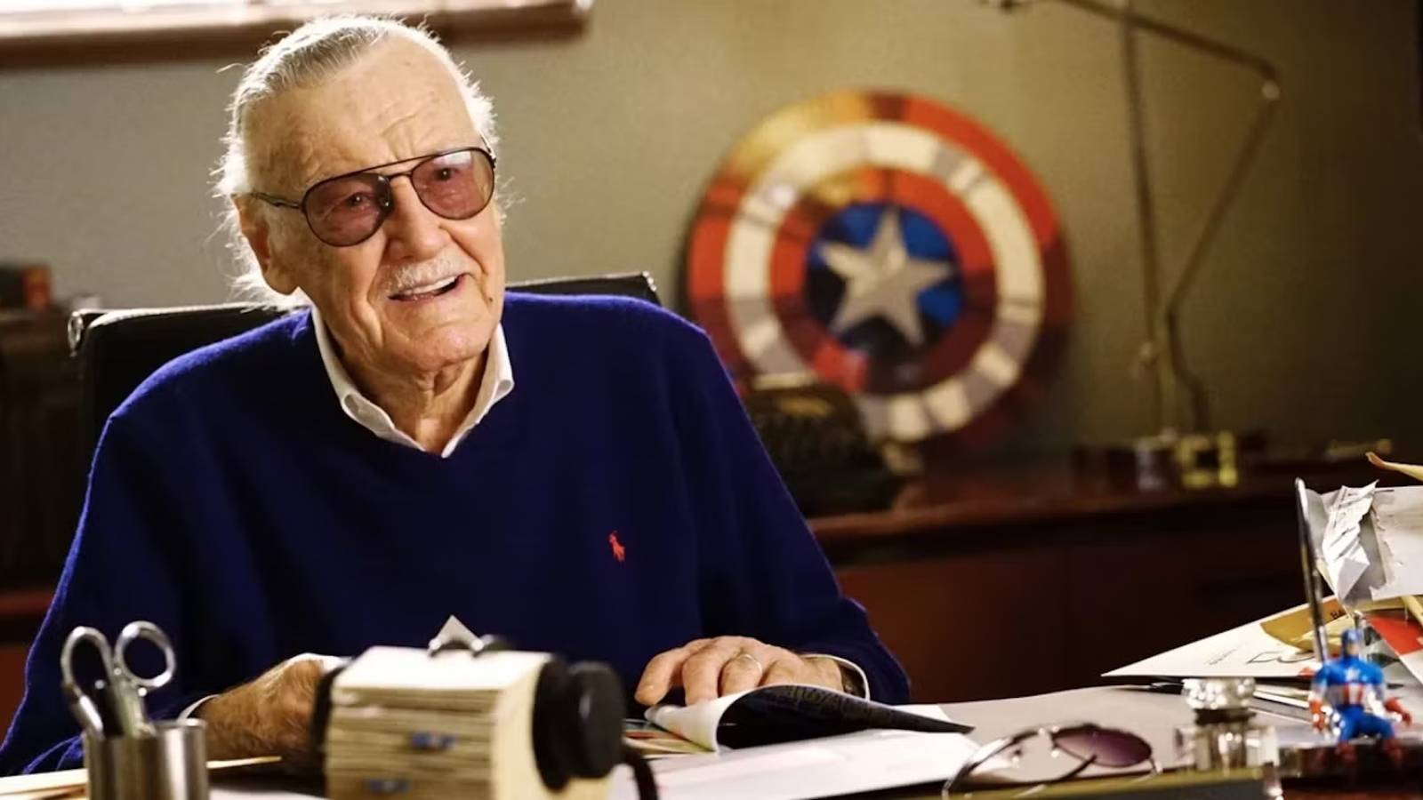 Stan Lee sitting at a desk with Captain America's shield in the background