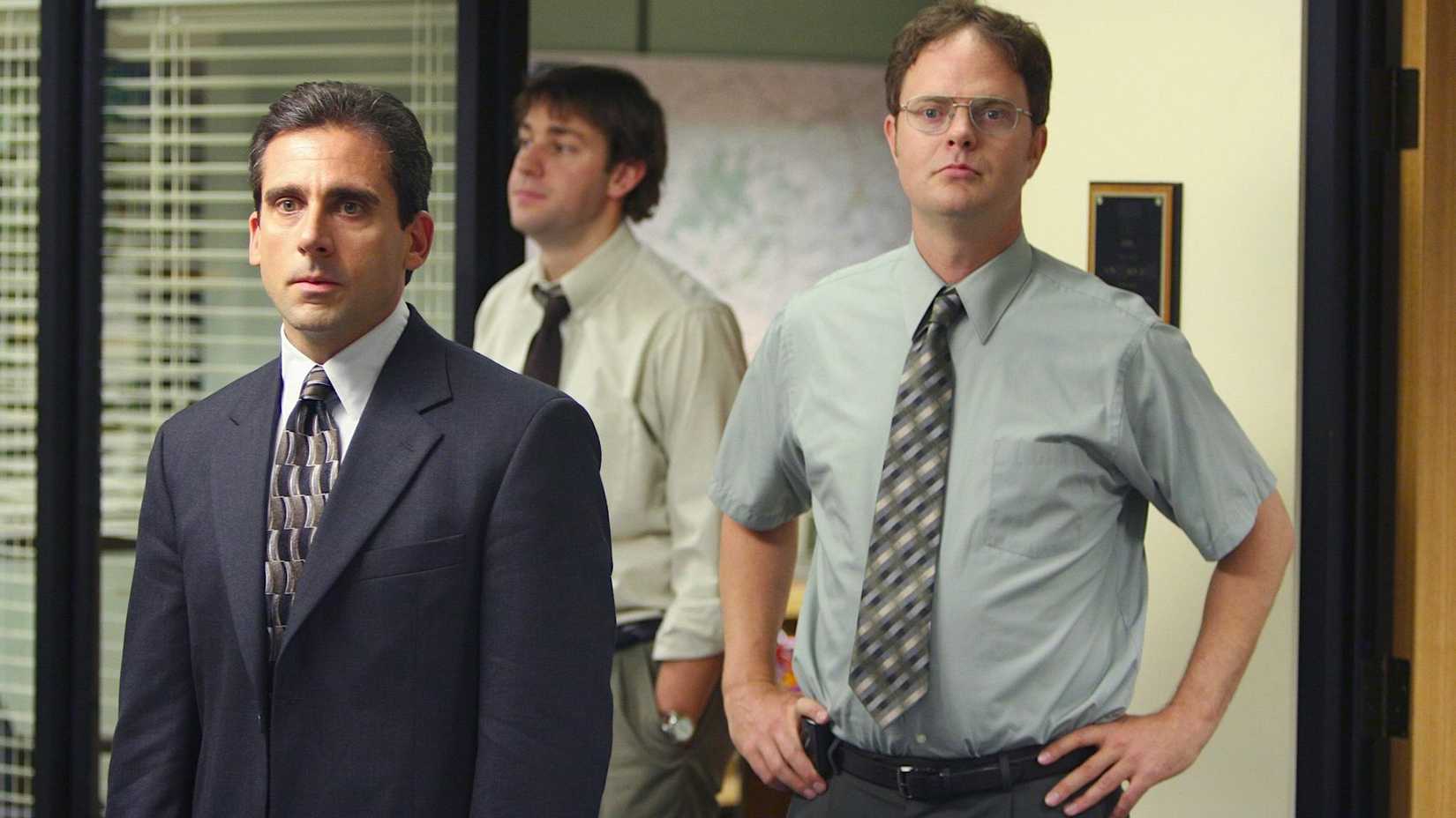 John Krasinski as Jim stepping out of a room behind Steve Carell as Michael and and Rainn Wilson as Dwight as they address the employees in The Office.