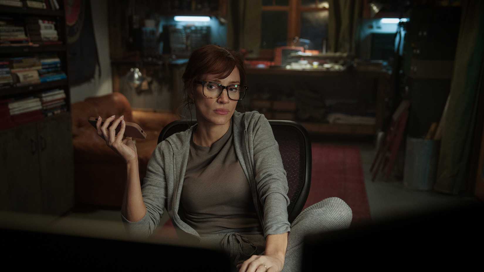 A woman sitting in front of a computer holding out a phone in The Savant.
