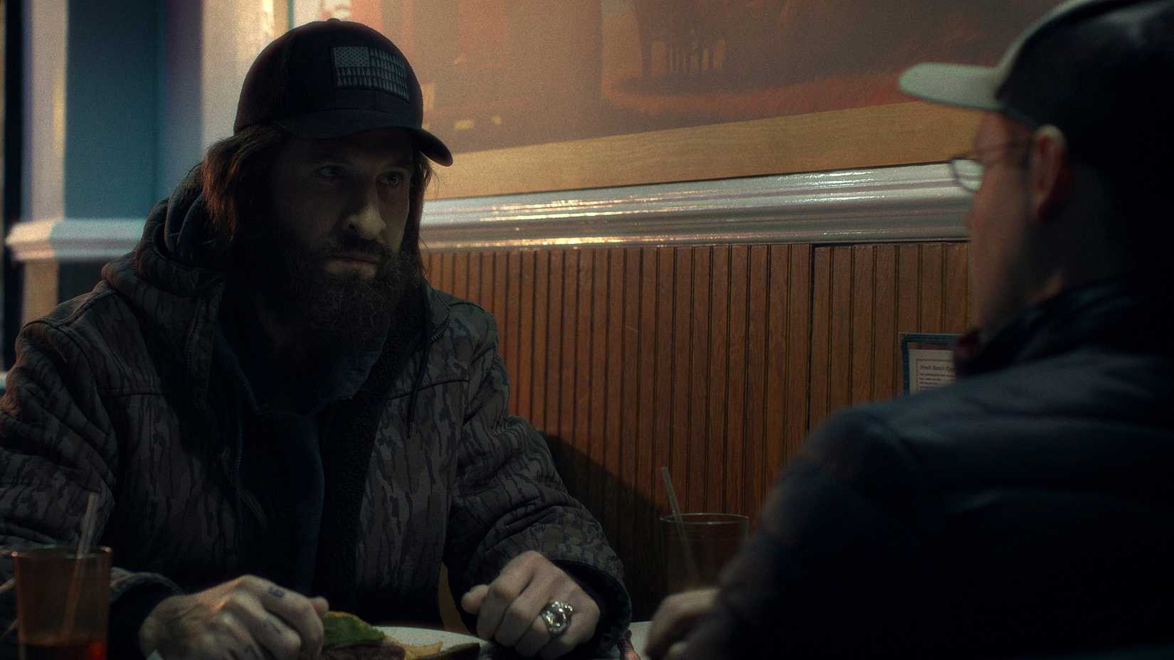 A scruffy man sitting in a diner across from another in The Savant.