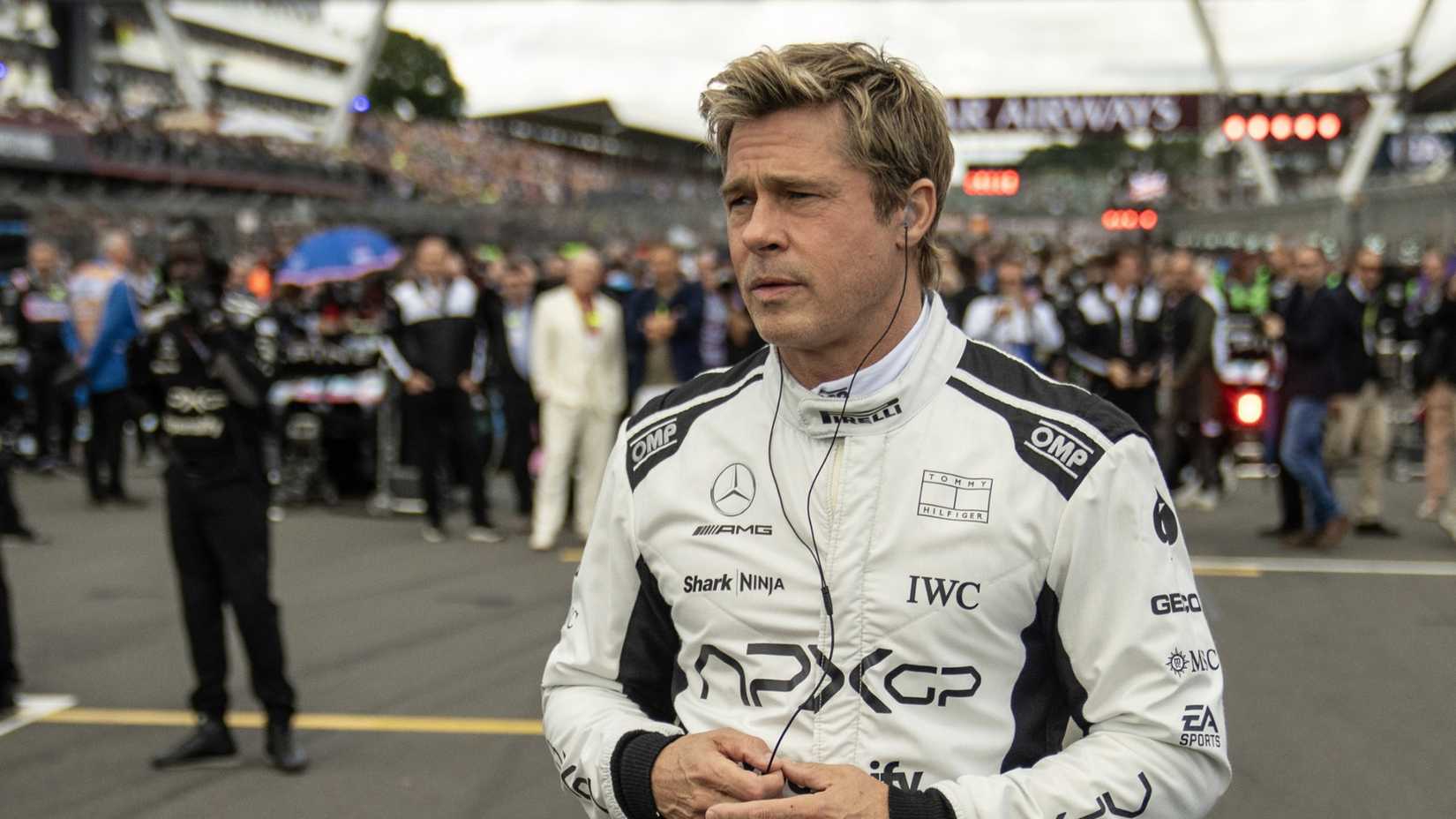 Brad Pitt in F1 wearing his uniform, standing on the track with headphones in