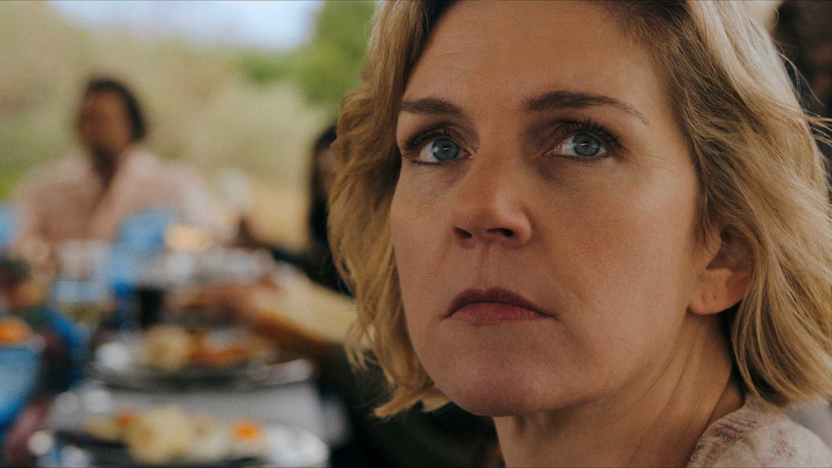 A close-up of Carol looking behind her while at a table with others in Pluribus.