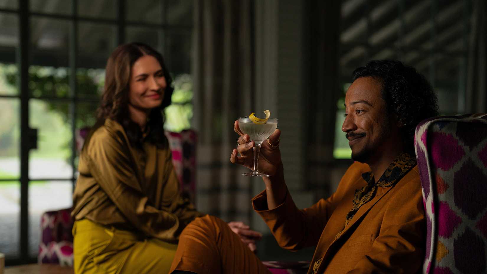 Zosia sitting with Kumba, who is enjoying a martini in Pluribus.