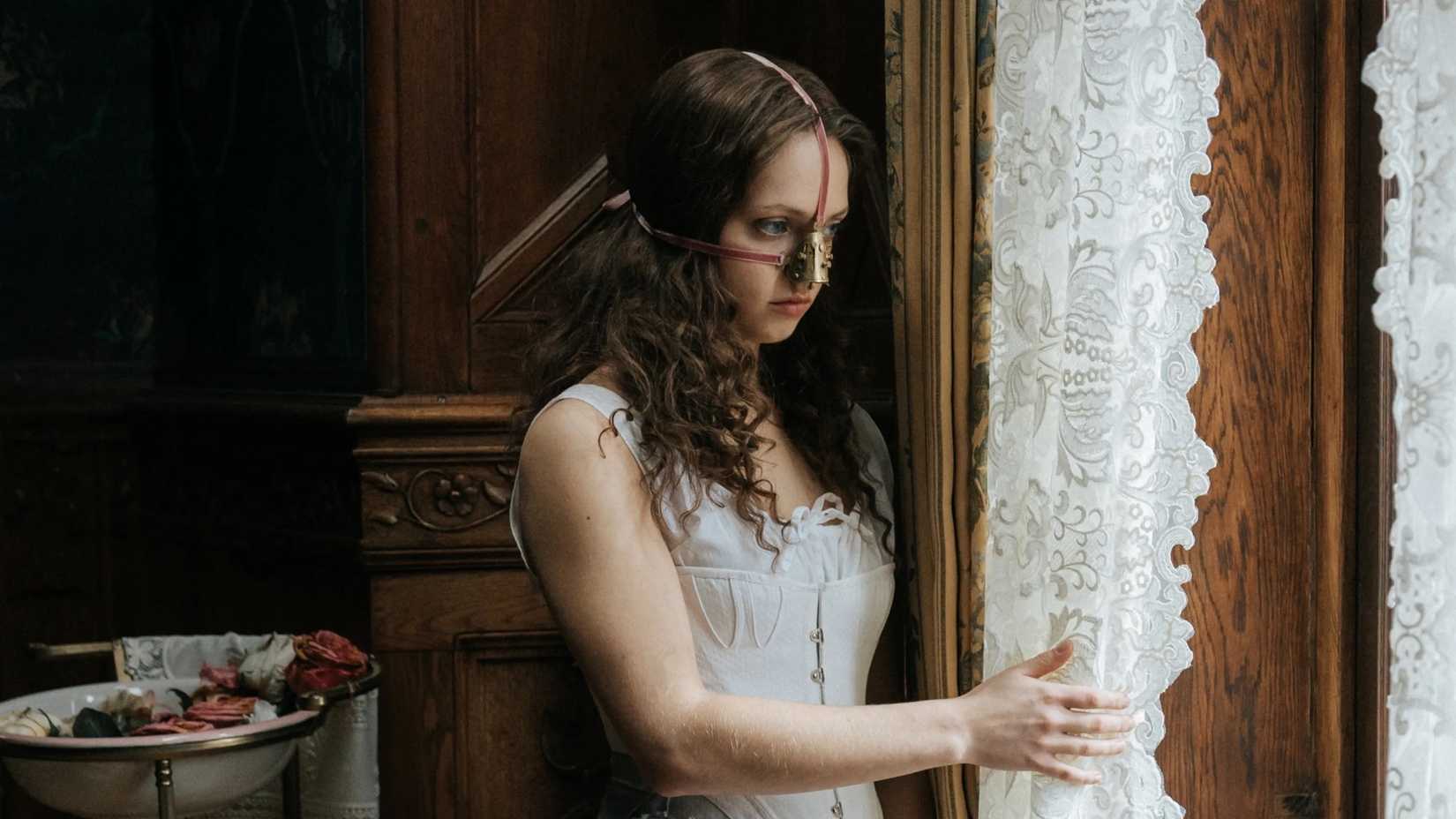 Seorang wanita dengan alat logam di wajahnya melihat ke luar jendela dalam film The Ugly Stepsister.
