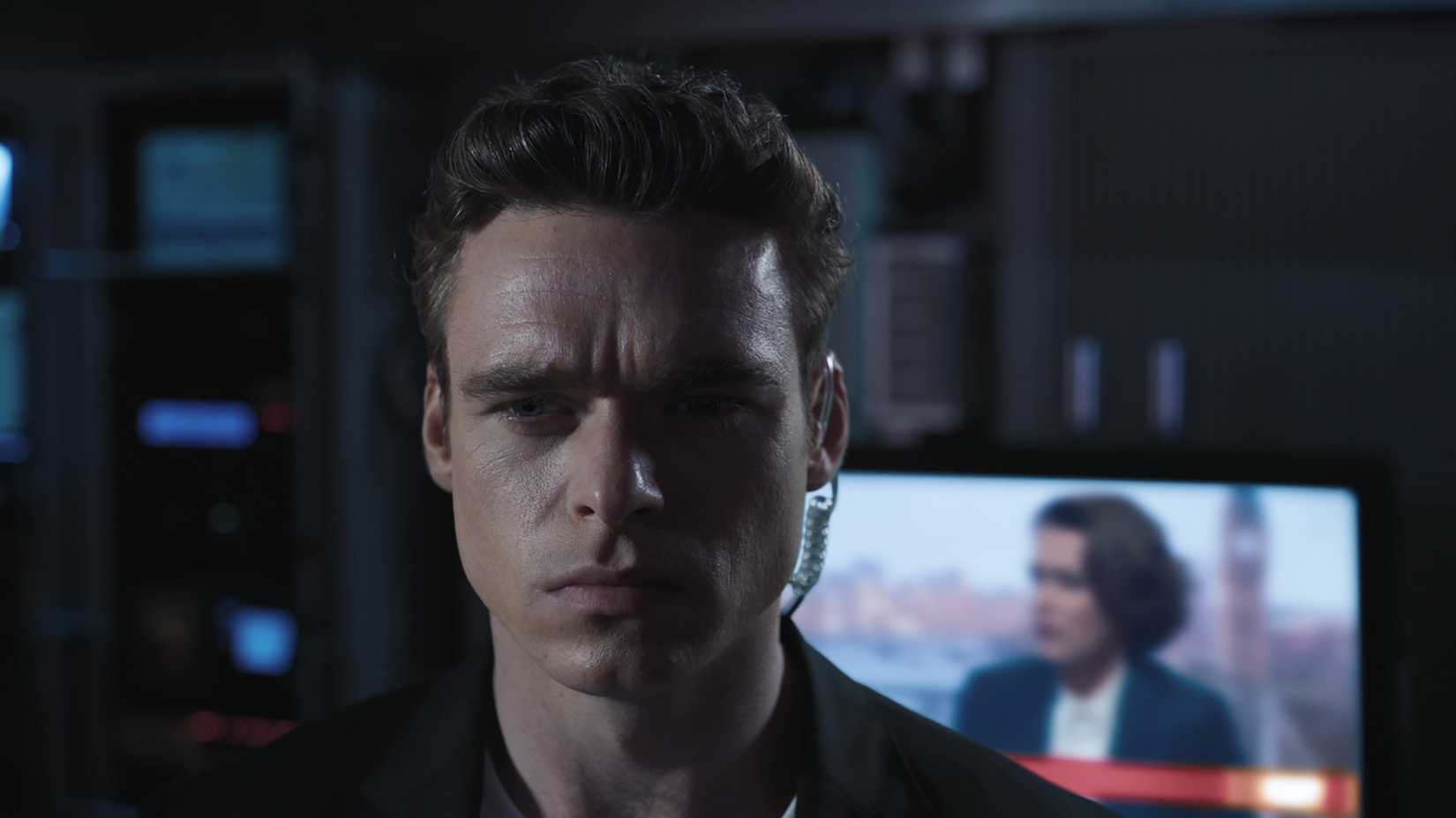A close-up of David looking angry, Julia on the TV in the background in Bodyguard.