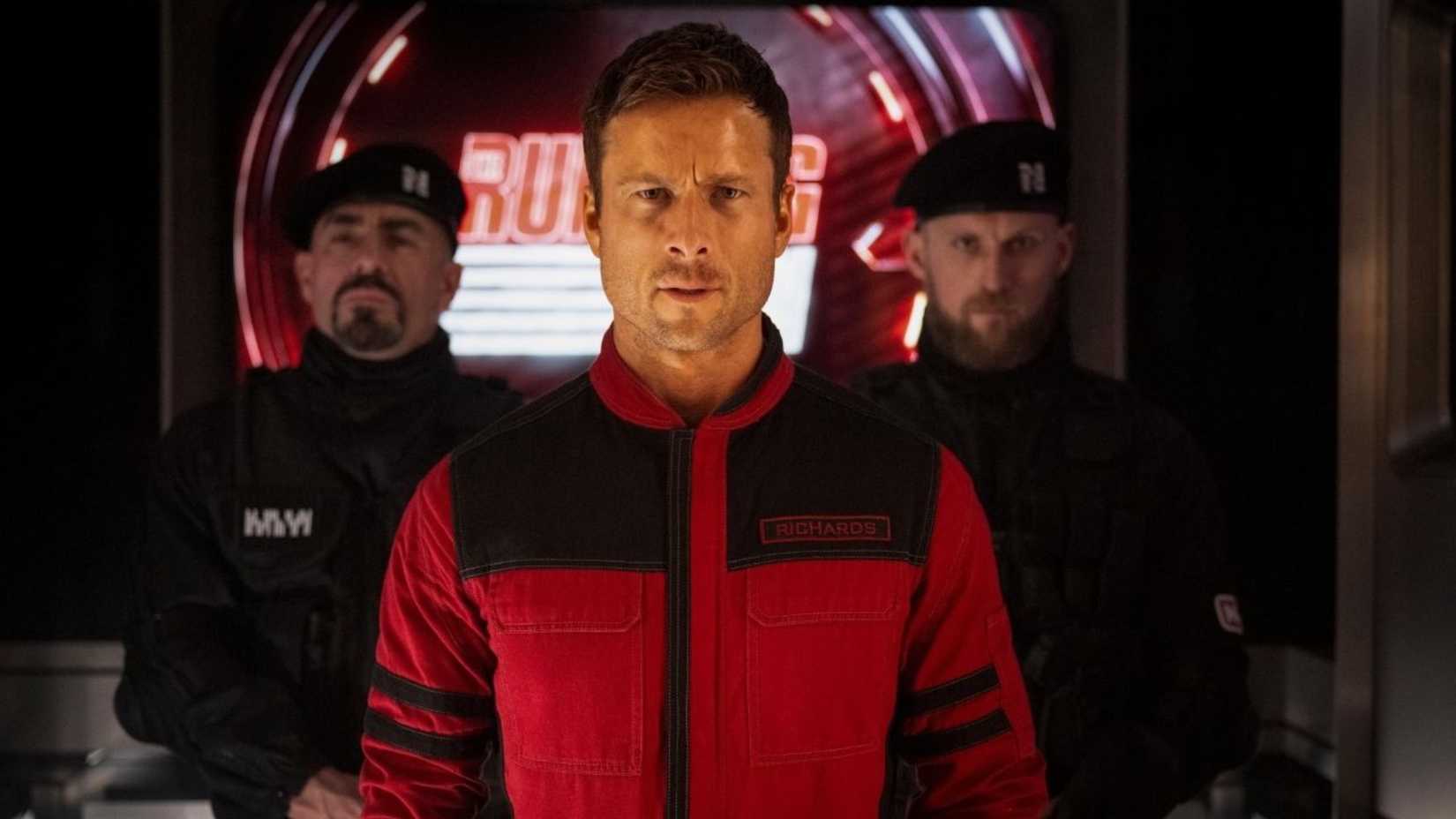 Glen Powell is escorted by two men in the film the running man