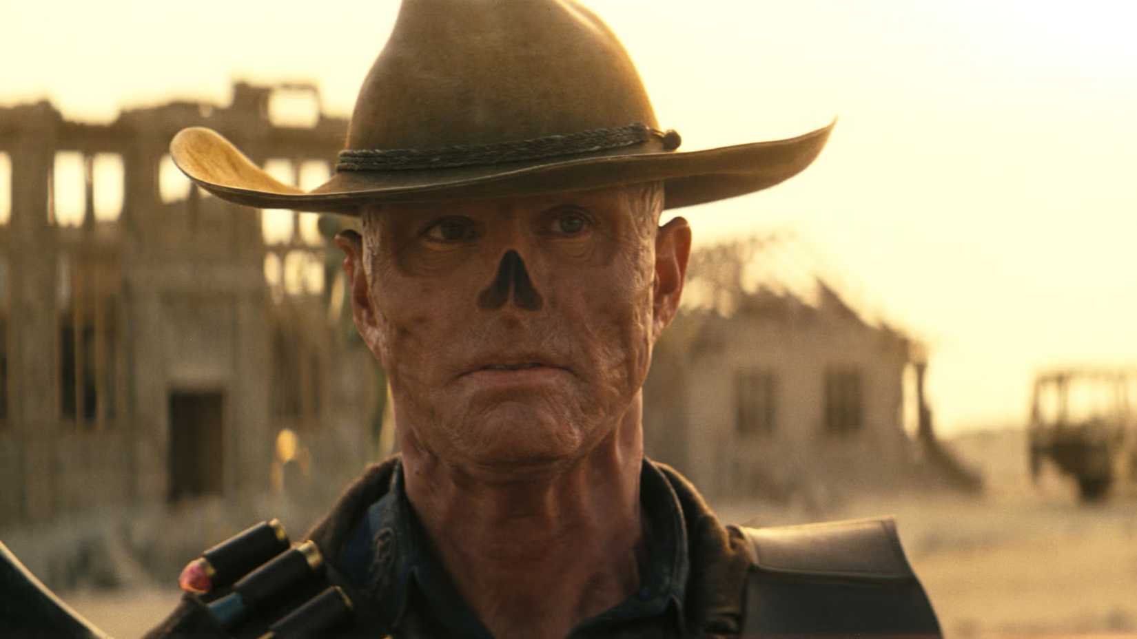 Walton Goggins as the Ghoul in Fallout wearing a cowboy hat with a broken down house behind him