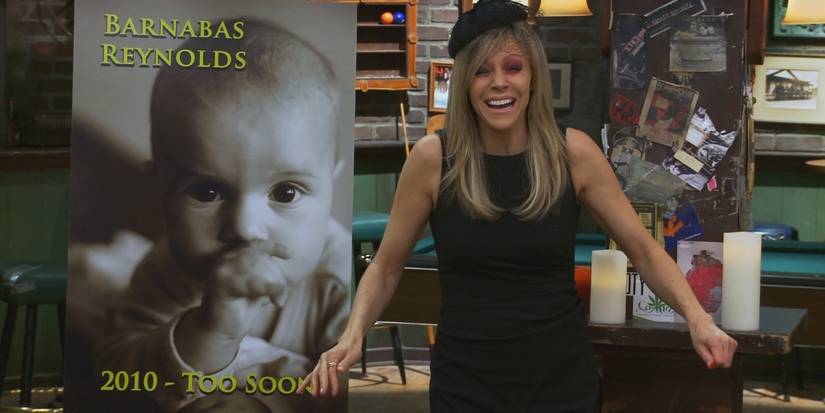 Dee crying at Paddy's Pub while standing in front of a sign for the fake funeral of baby Barnabas in It's Always Sunny in Philadelphia.