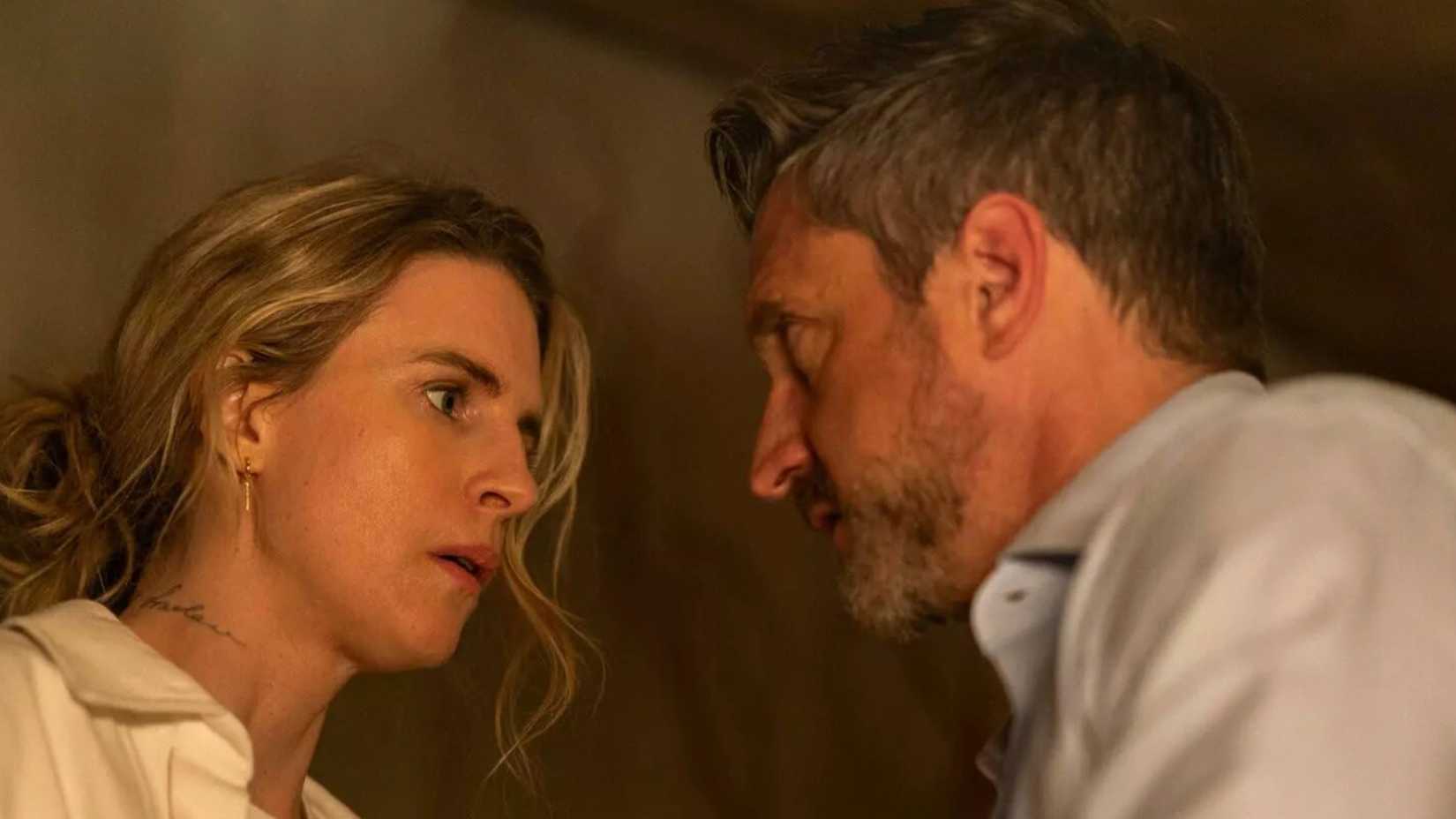 Brit Marling and Raúl Esparza staring seriously at each other in A Murder at the End of the World