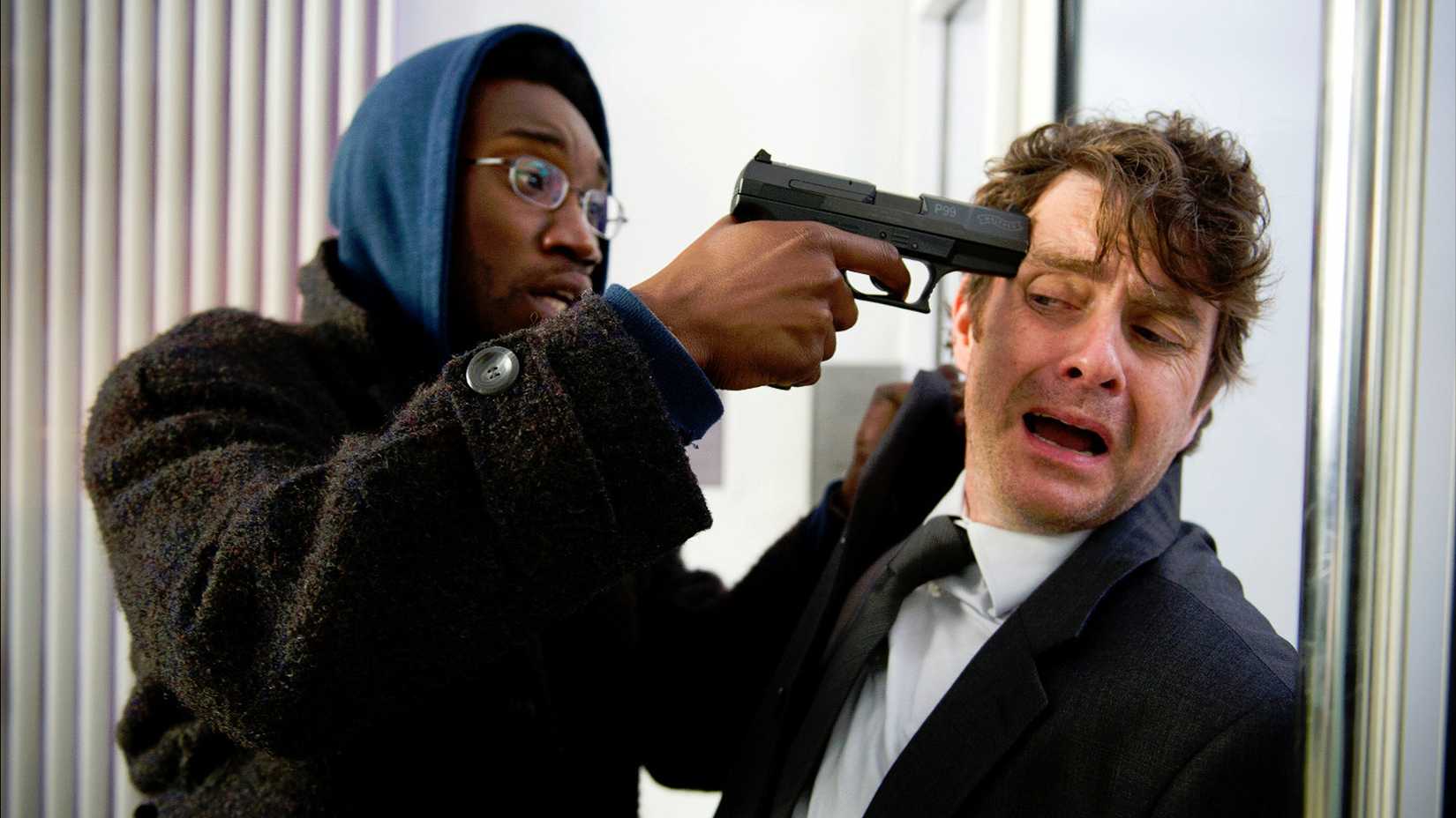 Michael looking terrified as Ian pushes him against a wall and holds a gun to his head in the UK version of Utopia.