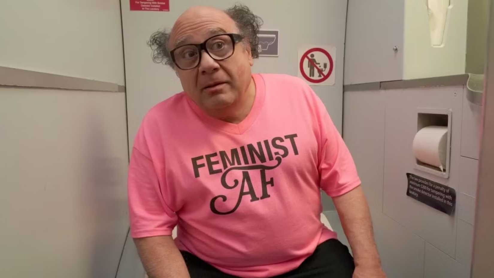 Danny DeVito as Frank Reynolds in sitting in an airplane bathroom wearing a pink feminist top in It's Always Sunny in Philadelphia.