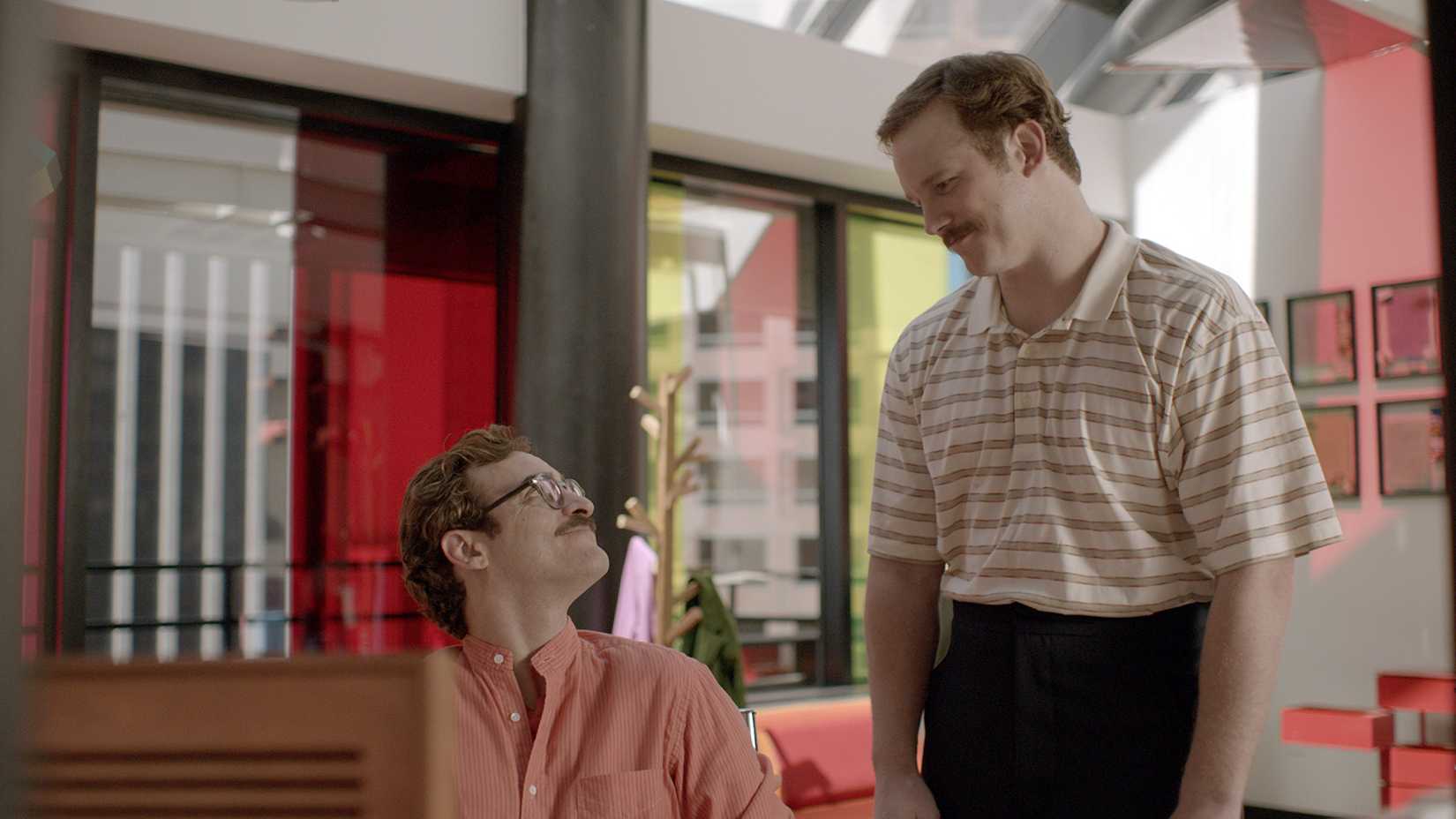 Theodore smiles back at Paul while sitting at his desk, chatting, in Her.