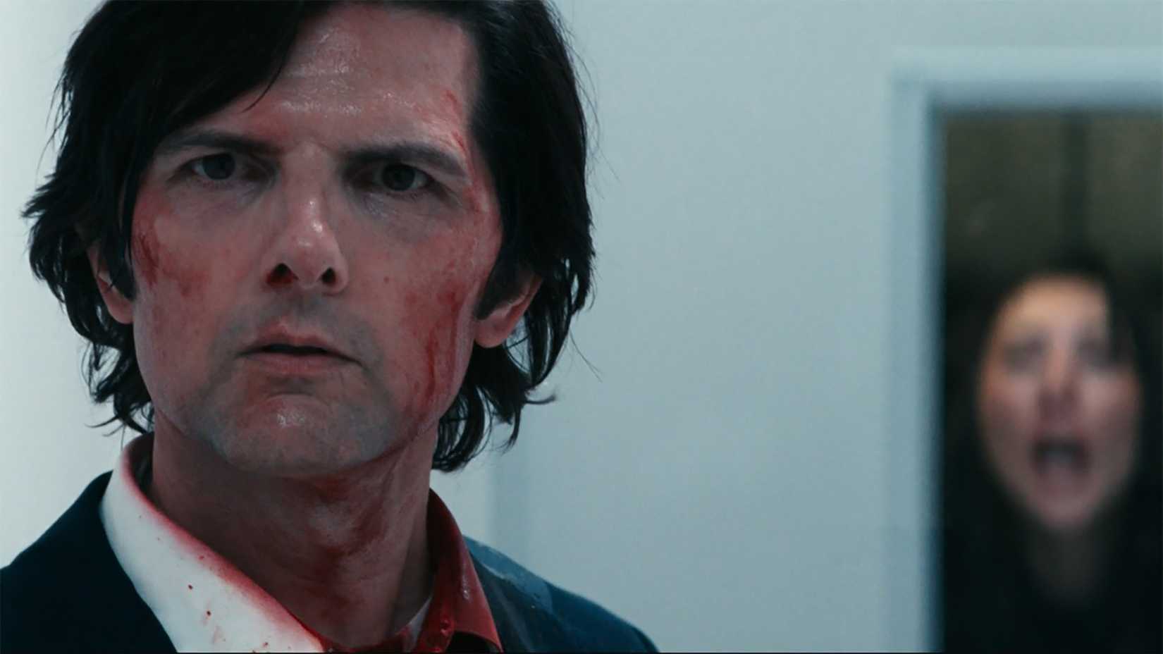 Mark with a bloody face staring intently in one direction, Gemma screaming behind a window behind him in Severance.