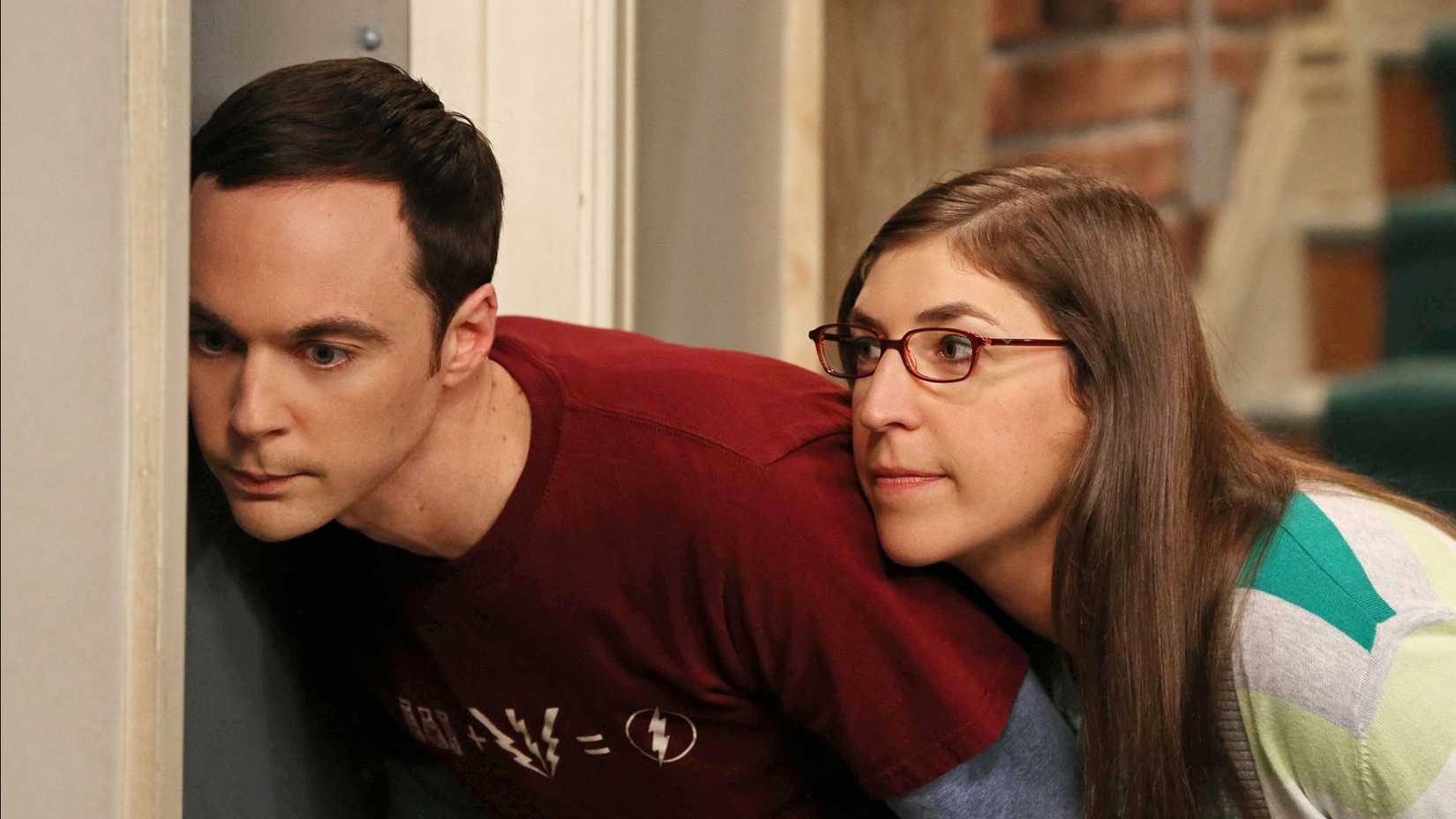 Amy leaning against Sheldon and watching him lovingly as he tries to listen through a door in The Big Bang Theory.