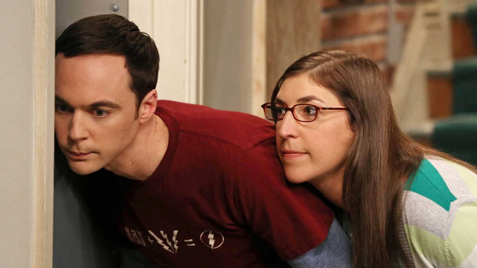 Amy leaning against Sheldon and watching him lovingly as he tries to listen through a door in The Big Bang Theory.