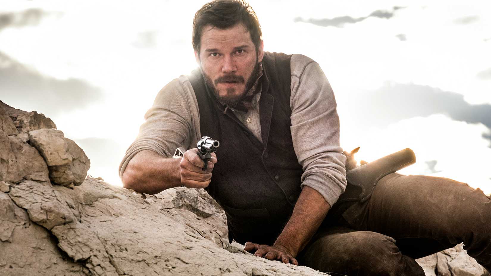 Chris Pratt as Joshua Faraday lying on a rock and pointing a gun forward in The Magnificent Seven.