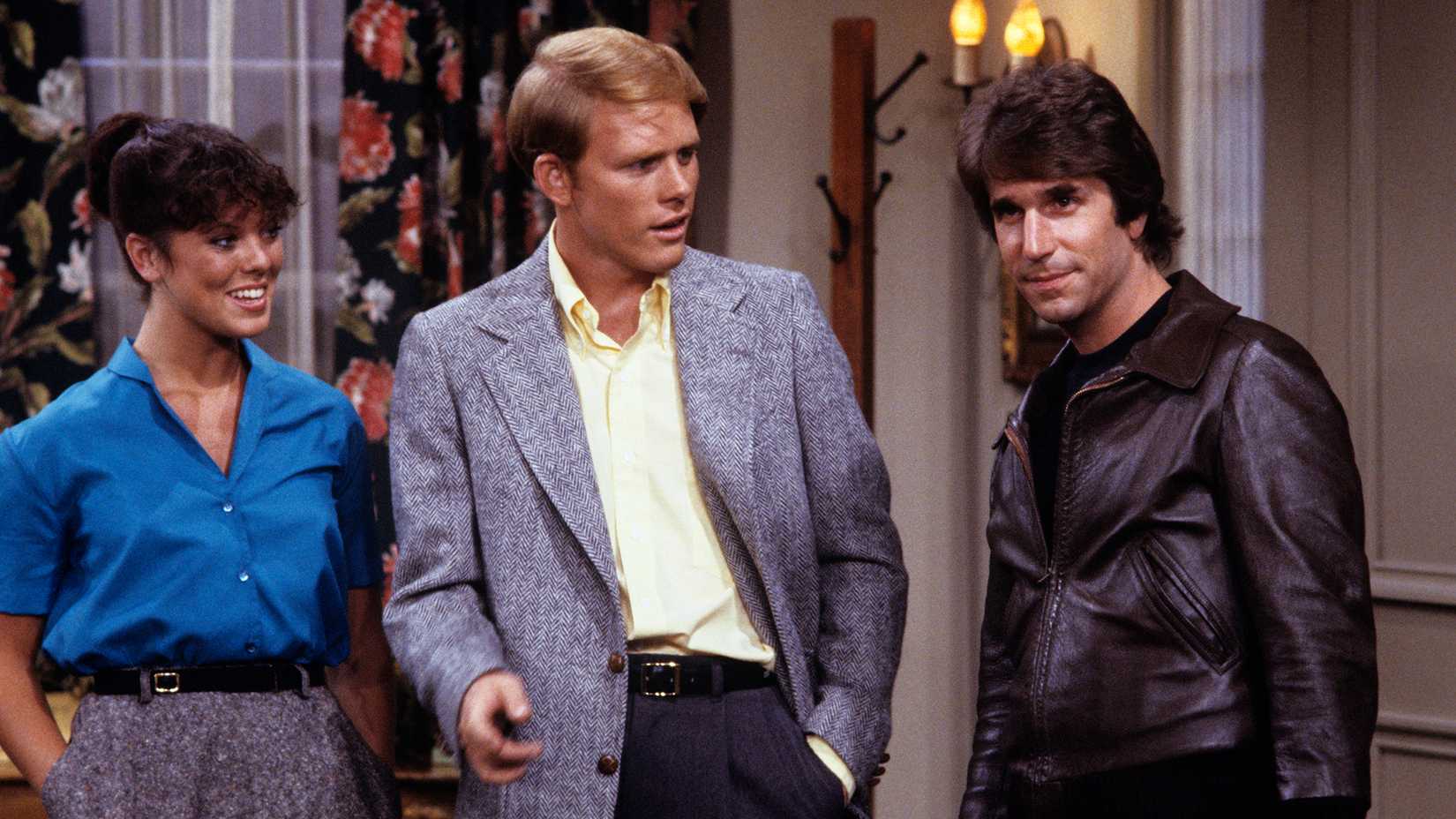 Richie and Joanie watching a smirking Fonzie while standing together in the hallway in Happy Days.