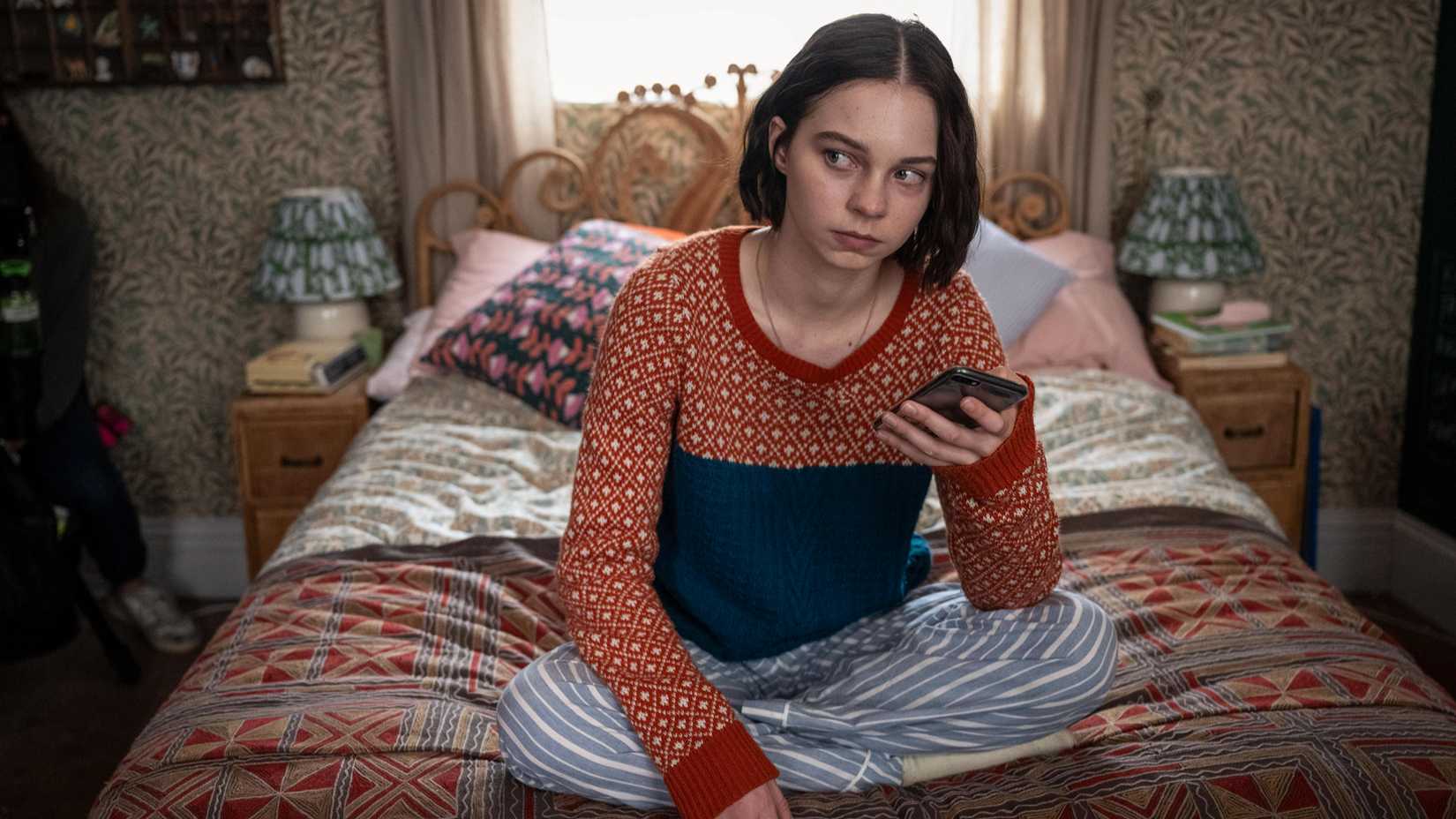 Pip sitting on her bed cross legged, holding her phone and staring off at something, in A Good Girl's Guide to Murder.