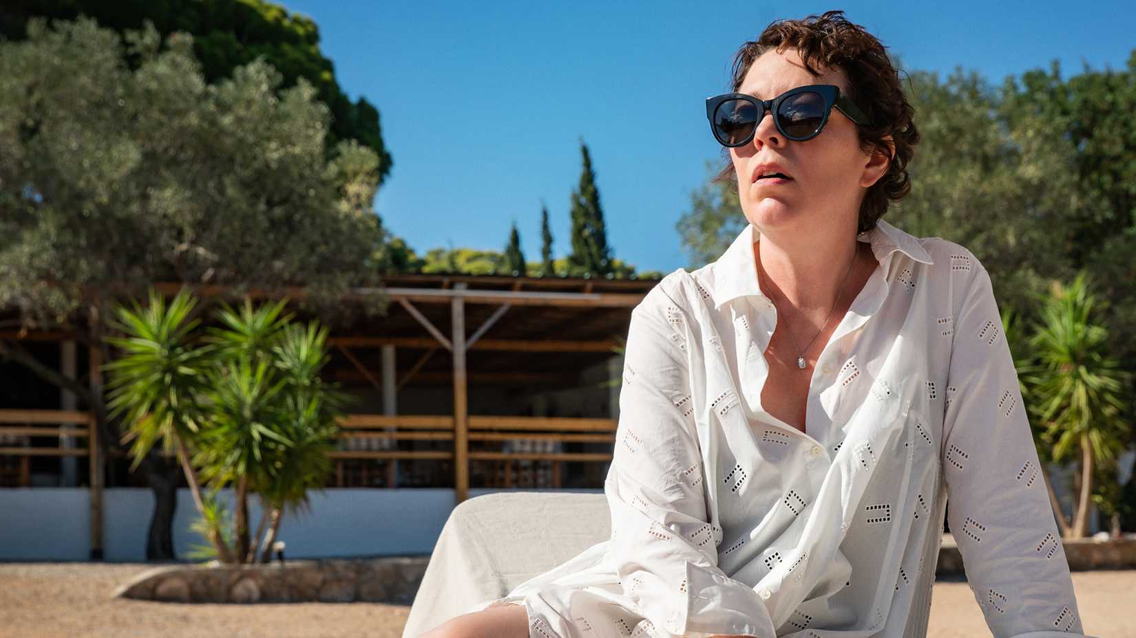 Olivia Colman as Leda wearing sunglasses as she lays in the sun, looking concerned at something, in The Lost Daughter.