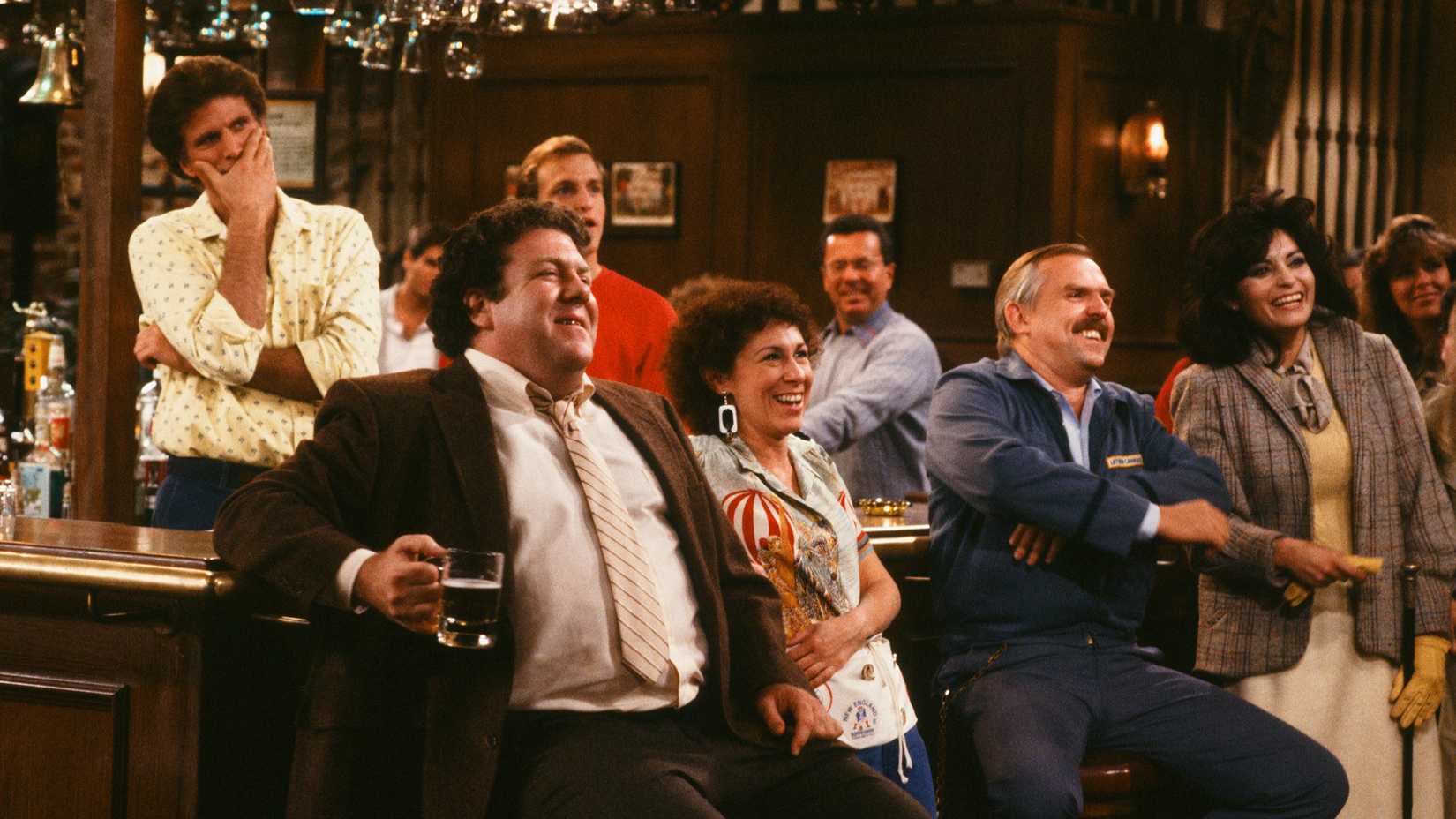 Sam, Norm, Woody, Carla, and Cliff sat around the bar, watching something off-screen, in Cheers.