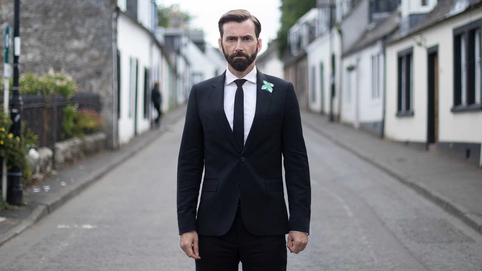 David Tennant as Tom Kendrick, standing in the middle of a street while wearing a black suit, in Deadwater Fell.