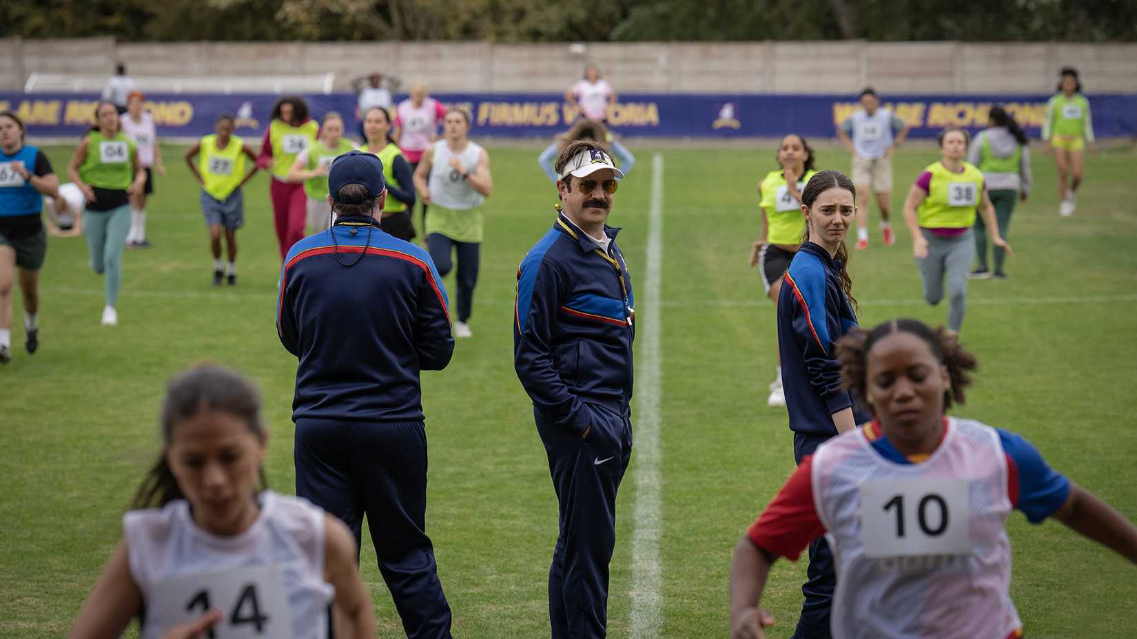Ted standing in the middle of the field with Alice as the women practice in Ted Lasso.