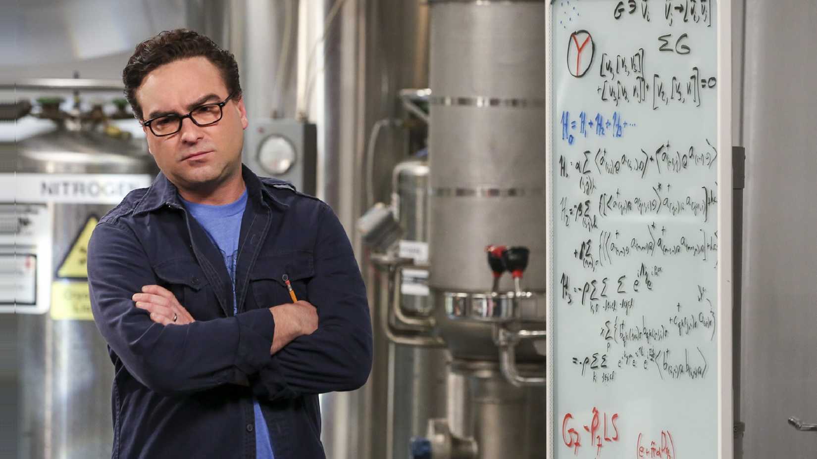 Johnny Galecki as Leonard Hofstadter with his arms crossed, standing next to a whiteboard with equations on it, in The Big Bang Theory.
