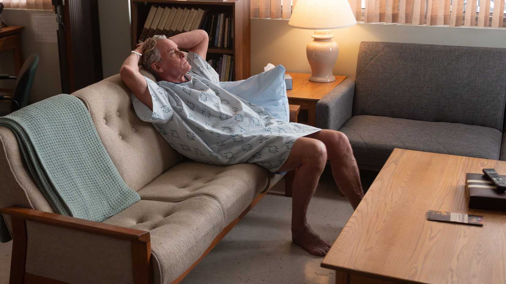 John C. McGinley as Dr. Cox sitting on a coach in a hospital gown in Scrubs